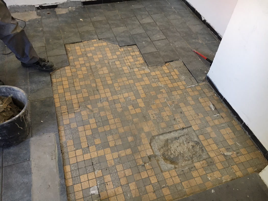 UZIN_Application_Image_UZIN NC 110_0445_ 2018-03 Djelomično uklonjena pločica poda tijekom renovacije: u prvom planu male bež‑sive mozaik pločice s istrošenim i nedostajućim dijelovima; veće tamne pravokutne pločice uz rub podignute su, otkrivajući ljepilo i podlogu. Lijevo se vide noge i crne radne čizme radnika pored kante s uklonjenim pločicama; desno se crvena poluga naslanja na zid. Bijeli zidovi s crnom lajsnom, prašina i krhotine razasute po podu. Fotografija snimljena iz niskog kuta gledano prema dolje.
