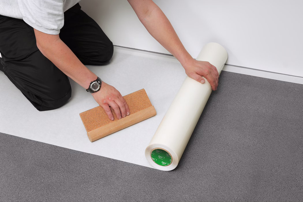 UZIN_Application Image_Sigan1_1_2019-05 A worker kneels indoors installing flooring underlayment along a white wall. Only the person's forearms and hands are visible; one hand is unrolling a wide white underlayment (foam or vapor barrier) while the other holds a wooden smoothing tool with a cork surface. A roll of underlayment rests on a gray vinyl or linoleum floor; the worker wears black work trousers, a white T-shirt and a black wristwatch. The scene is well lit and focused on the flooring installation process and tools.